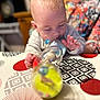 adult, baby, blurred_background, bottle, child, clothing, colorful, curious, exploration, face, focus, hand, head, indoor, pattern, person, playing, skin, tablecloth, toy