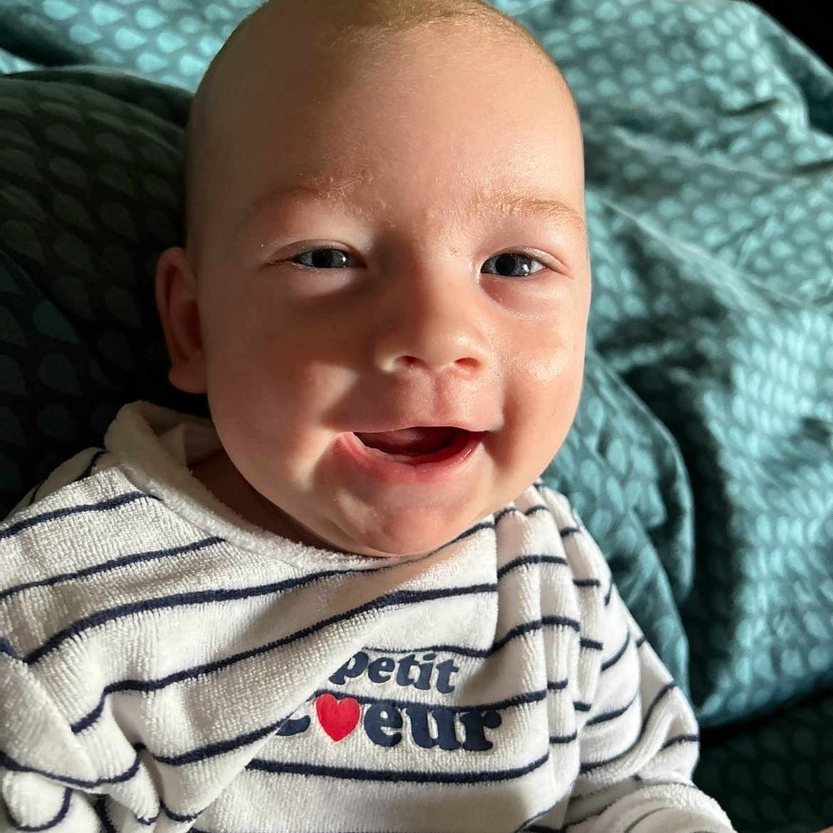 Djuliane participe au concours pour gagner de l'argent avec cette photo : baby, bed, blanket, blue, child, clothing, cozy, cute, face, happy, head, indoor, infant, person, portrait, skin, smile, striped_shirt, text_on_clothing, young