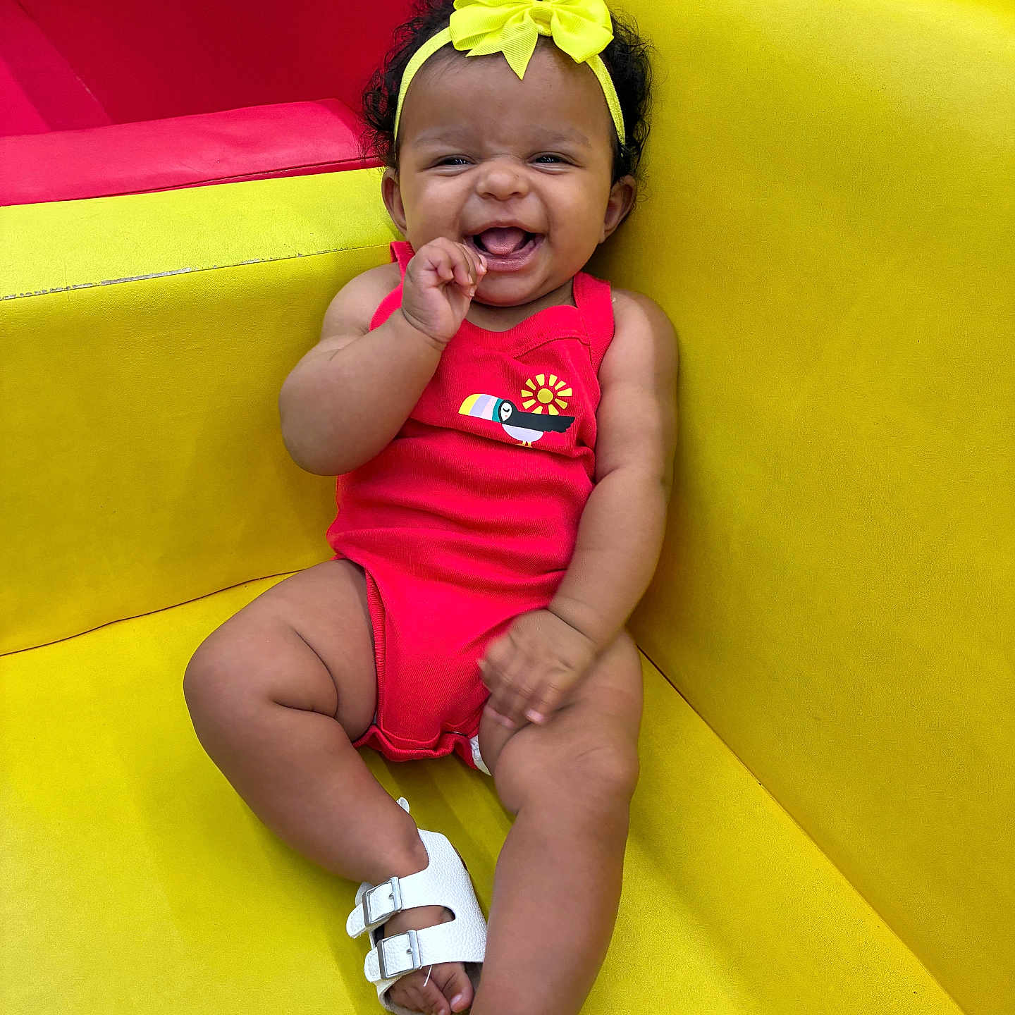 Shanyla is registered to the contest to win money with this photo: baby, bow, cheerful, child, colorful, curly_hair, happy, headband, indoors, infant, pink, playroom, portrait, red, romper, sandals, sitting, smiling, toy_seating, yellow