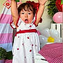 child, toddler, dress, strawberry_pattern, balloon, party, celebration, indoor, pink, red, white, plant, table, tablecloth, flower_balloon, curtain, smiling, happy, face, hands_up