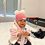 Louna participe au concours pour gagner de l'argent avec cette photo : baby, bed, black_pants, blurred_background, bottle, child, cozy_clothing, cute, home, indoor, person, pink_hat, pink_socks, pom_poms, sitting, smiling, toddler, vest, warm_clothes, window