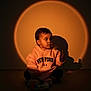 child, baby, hoodie, sitting, toy, rattle, shadow, spotlight, indoor, portrait, young_child, clothing, floor, expression, face, lighting, warm_tone, casual, cute, new_york