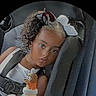 child, toddler, car_seat, car, passenger, portrait, face, eyes, hair_bows, curly_hair, smiling, serious_expression, sippy_cup, baby_bottle, seatbelt, travel, indoors, vehicle, closeup, photograph