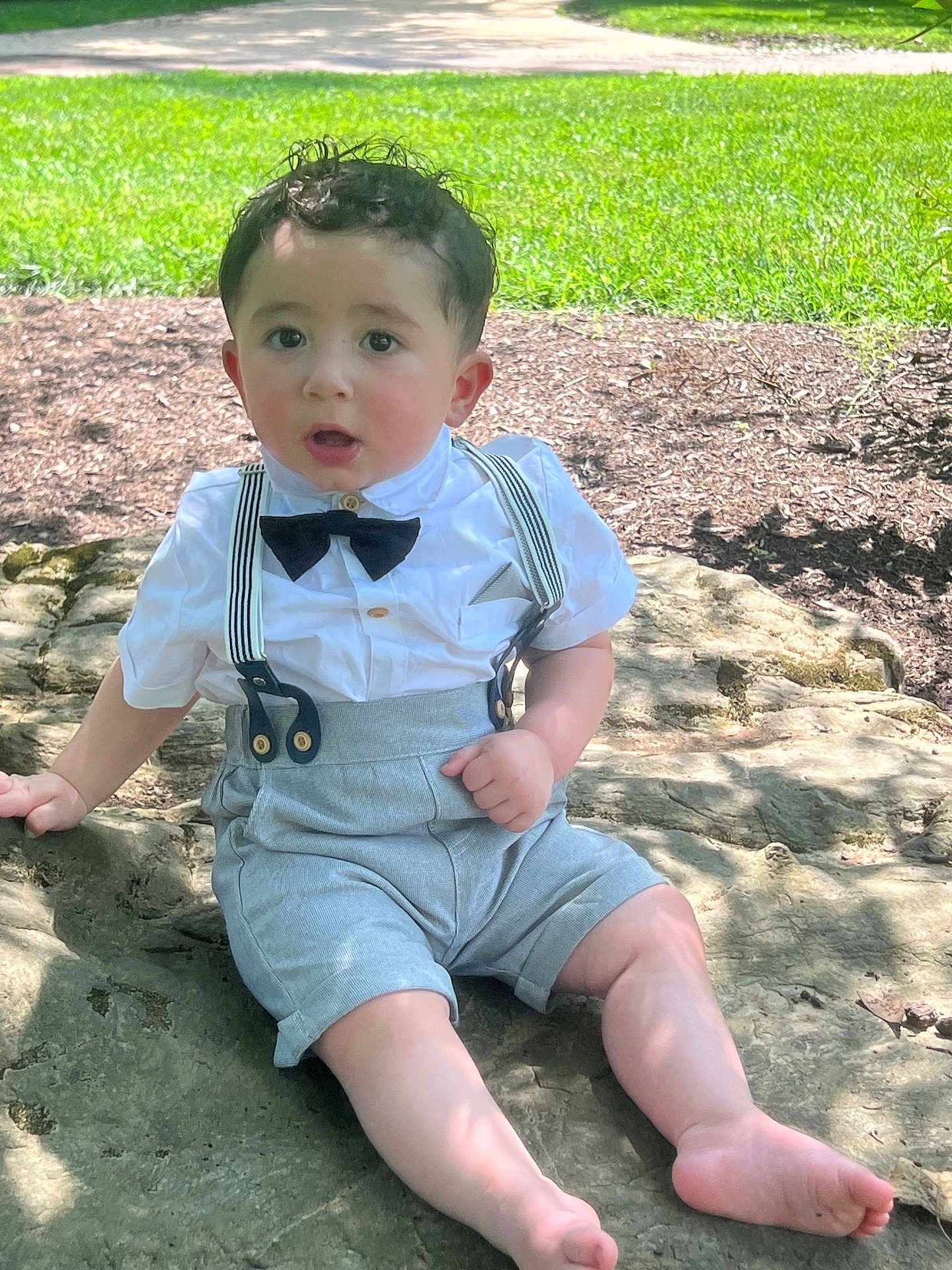 Oliver joined the competition — help win amazing prizes! toddler, child, boy, outdoor, grass, rock, sitting, clothing, white_shirt, bow_tie, suspenders, barefoot, face, curious, sunlight, nature, summer, portrait, cute, casual