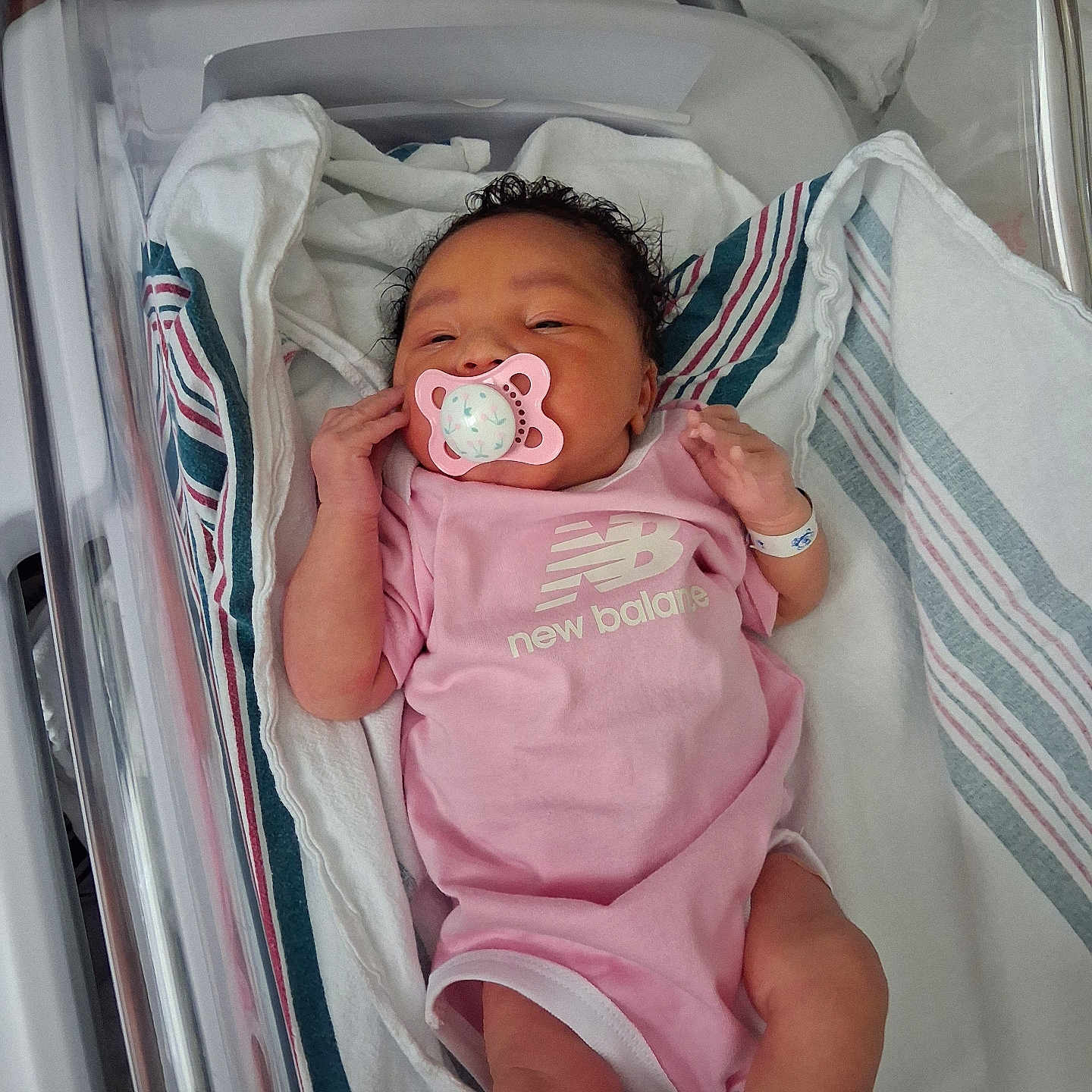 Aaliyah is registered to the contest to win money with this photo: newborn, baby, infant, hospital, bassinet, pacifier, pink_clothing, romper, blanket, swaddle, face, hair, hands, legs, sleeping, awake, portrait, medical_wristband, crib, new_balance