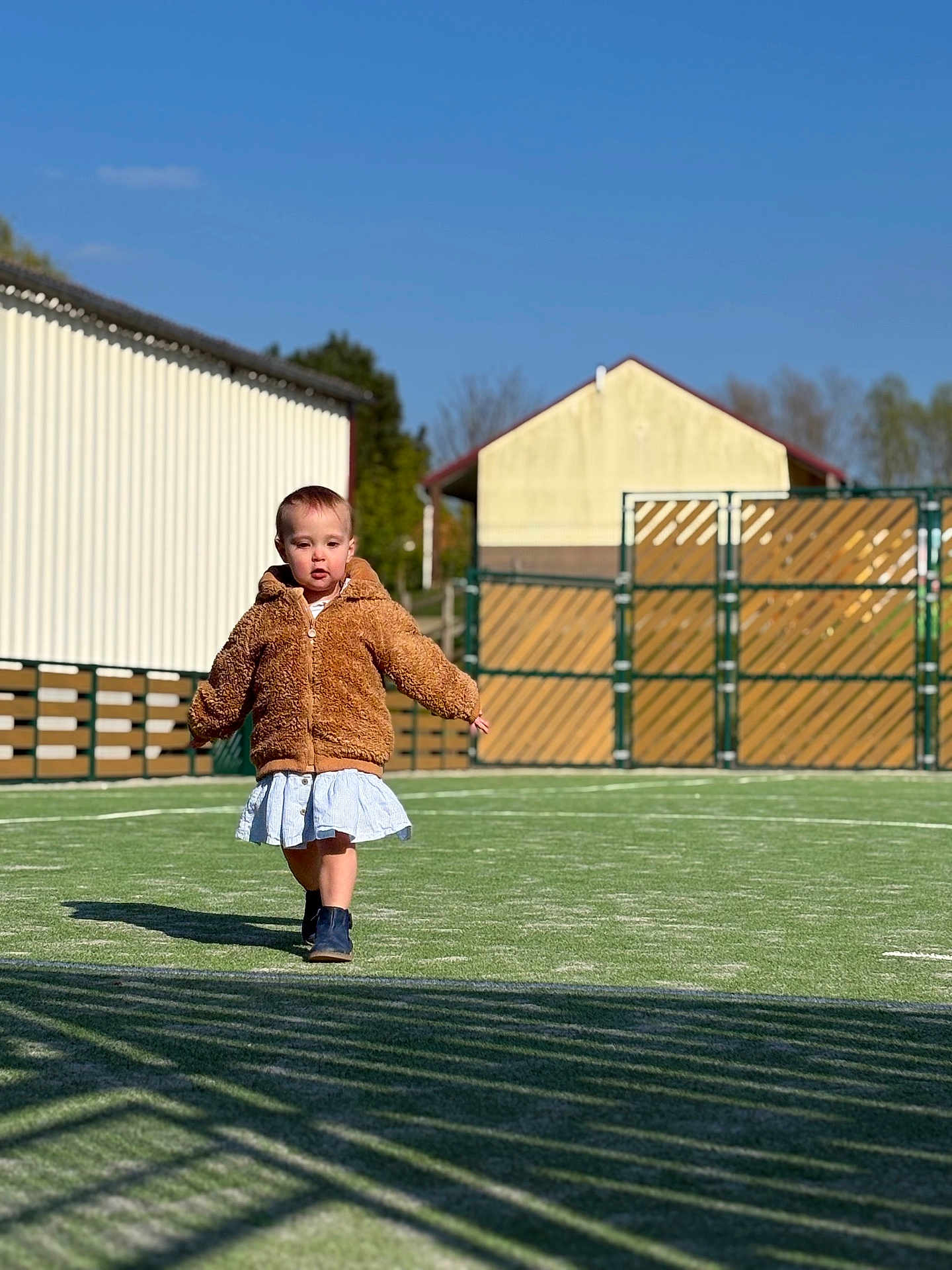 Iris participe au concours pour gagner de l'argent avec cette photo : child, toddler, walking, jacket, coat, dress, boots, astroturf, grass, playground, fence, building, sky, sunny, shadow, portrait, outdoor, daytime, person, play