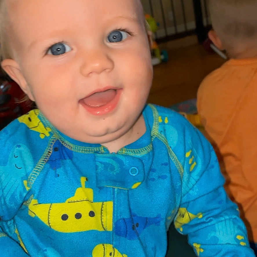 Carter is registered to the contest to win money with this photo: baby, child, blue_eyes, smiling, onesie, blue_clothing, indoor, happy, cute, person, infant, toddler, pattern, submarine, face, portrait, sitting, playroom, background, young_child