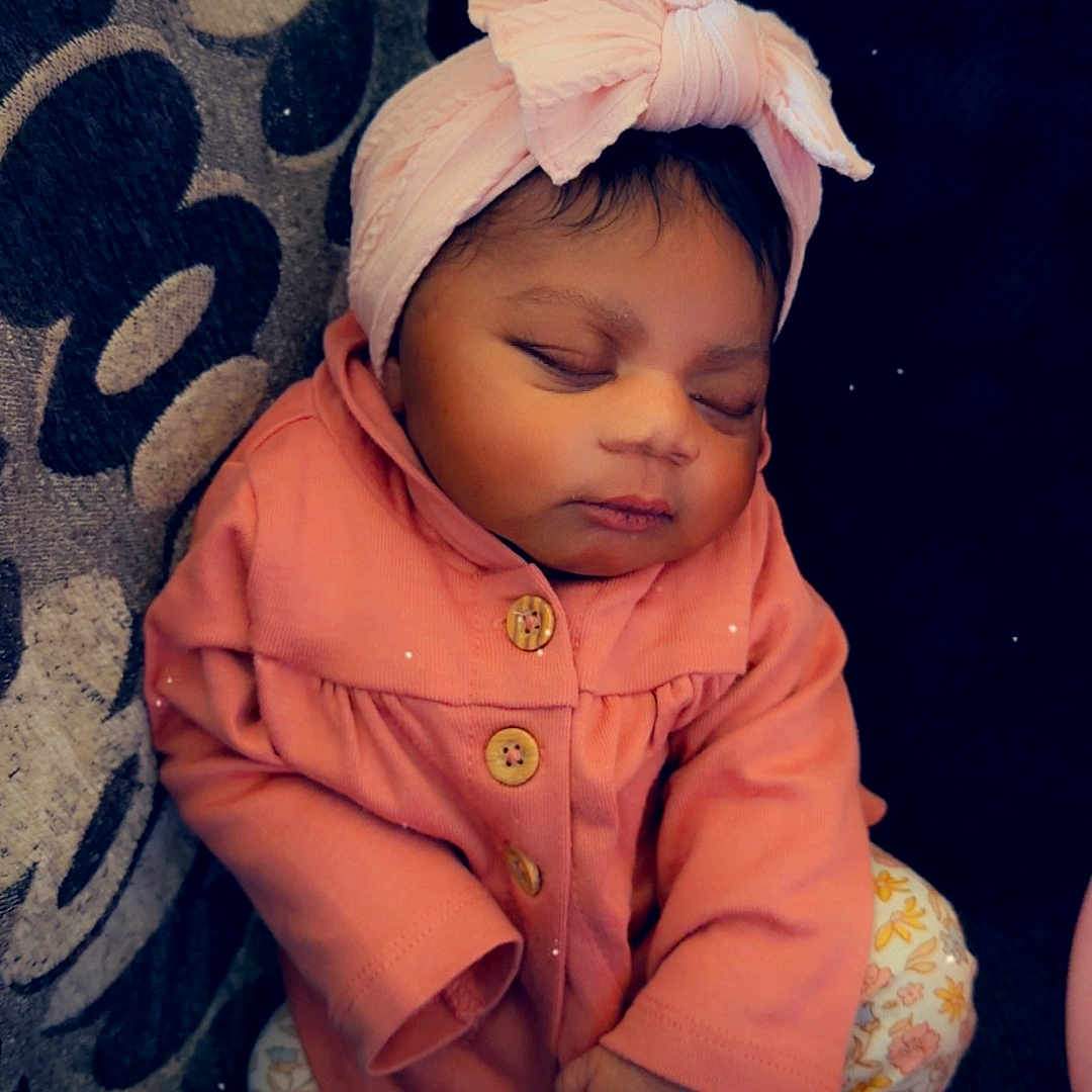 Na'Asia joined the competition — help win amazing prizes! baby, infant, sleeping, pink_jacket, headband, bow, couch, cushion, patterned_fabric, floral_pants, white_socks, peaceful, indoors, child, adorable, cute, resting, portrait, cozy, small_heart