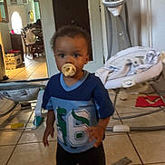 Julian is registered to the contest to win money with this photo: child, toddler, pacifier, blue_shirt, indoor, living_room, baby_chair, tile_floor, furniture, toy, door, blanket, baby_equipment, curly_hair, person, floor, home, casual_clothing, baby, cute