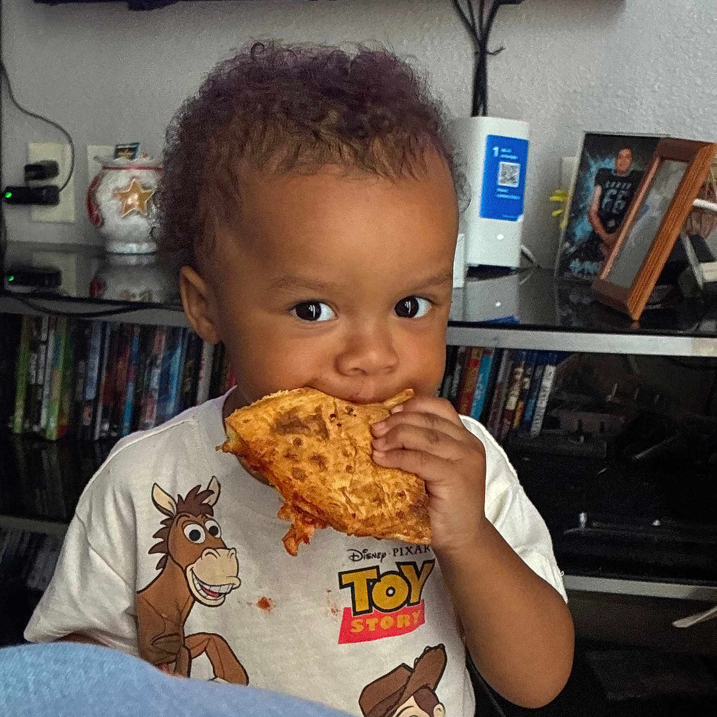 Julian is registered to the contest to win money with this photo: toddler, child, pizza, food, eating, curly_hair, toy_story, tshirt, indoor, living_room, television, shelf, photograph, frame, furniture, person, cute, snack, young_child, home