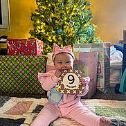 Shiloh is registered to the contest to win money with this photo: baby, child, pink_outfit, headband, milestone_card, 9_months, christmas_tree, christmas_lights, presents, gift_wrap, quilt, floor, holiday, festive, indoors, smiling, sitting, cute, happy, celebration