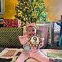 baby, child, pink_outfit, headband, milestone_card, 9_months, christmas_tree, christmas_lights, presents, gift_wrap, quilt, floor, holiday, festive, indoors, smiling, sitting, cute, happy, celebration