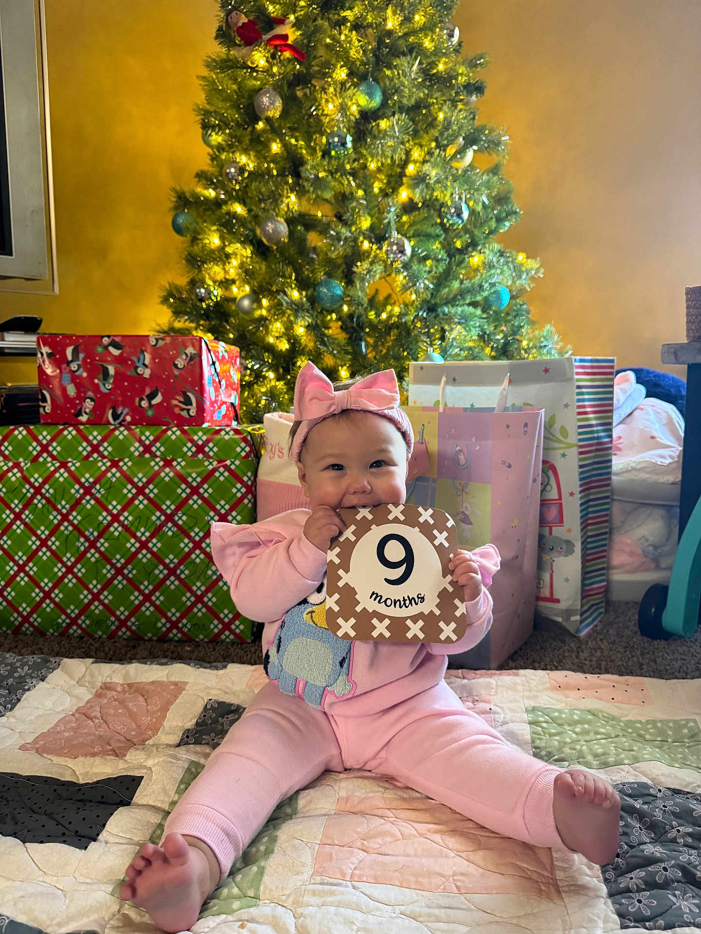 Shiloh is registered to the contest to win money with this photo: baby, child, pink_outfit, headband, milestone_card, 9_months, christmas_tree, christmas_lights, presents, gift_wrap, quilt, floor, holiday, festive, indoors, smiling, sitting, cute, happy, celebration