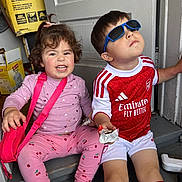 Angela Ademi is registered to the contest to win money with this photo: child, children, sitting, pink_clothing, sunglasses, red_jersey, snack, smiling, curly_hair, short_hair, indoor, steps, door, bag, footwear, happy, casual, playful, toddler, cute