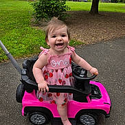 Remington is registered to the contest to win money with this photo: baby, child, toddler, smiling, happy, outdoors, park, toy_car, pink, dress, strawberry, steering_wheel, vehicle, playtime, standing, infant, portrait, grass, trees, road