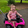 baby, child, toddler, smiling, happy, outdoors, park, toy_car, pink, dress, strawberry, steering_wheel, vehicle, playtime, standing, infant, portrait, grass, trees, road