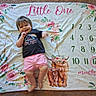 Remington is registered to the contest to win money with this photo: baby, child, toddler, smiling, lying_down, portrait, milestone_blanket, floral_blanket, pink_shorts, black_shirt, pink_floyd_shirt, carpet, indoor, numbers, highland_cow, flowers, roses, greenery, happy, cozy