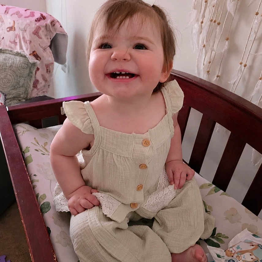 Remington is registered to the contest to win money with this photo: baby, child, infant, smiling, happy, portrait, sitting, crib, nursery, bow, romper, dress, clothing, brown_hair, face, teeth, indoor, bedding, curtain, cozy