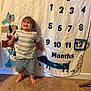 toddler, child, smiling, standing, barefoot, carpet, milestone_blanket, numbers, dogs, striped_shirt, shorts, indoor, happy, cute, young_child, wall, fabric, playful, portrait, home