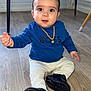 baby, child, floor, blue_shirt, white_pants, black_shoes, necklace, indoor, wooden_floor, cute, sitting, person, young_child, expression, looking, footwear, flooring, home, furniture, legs