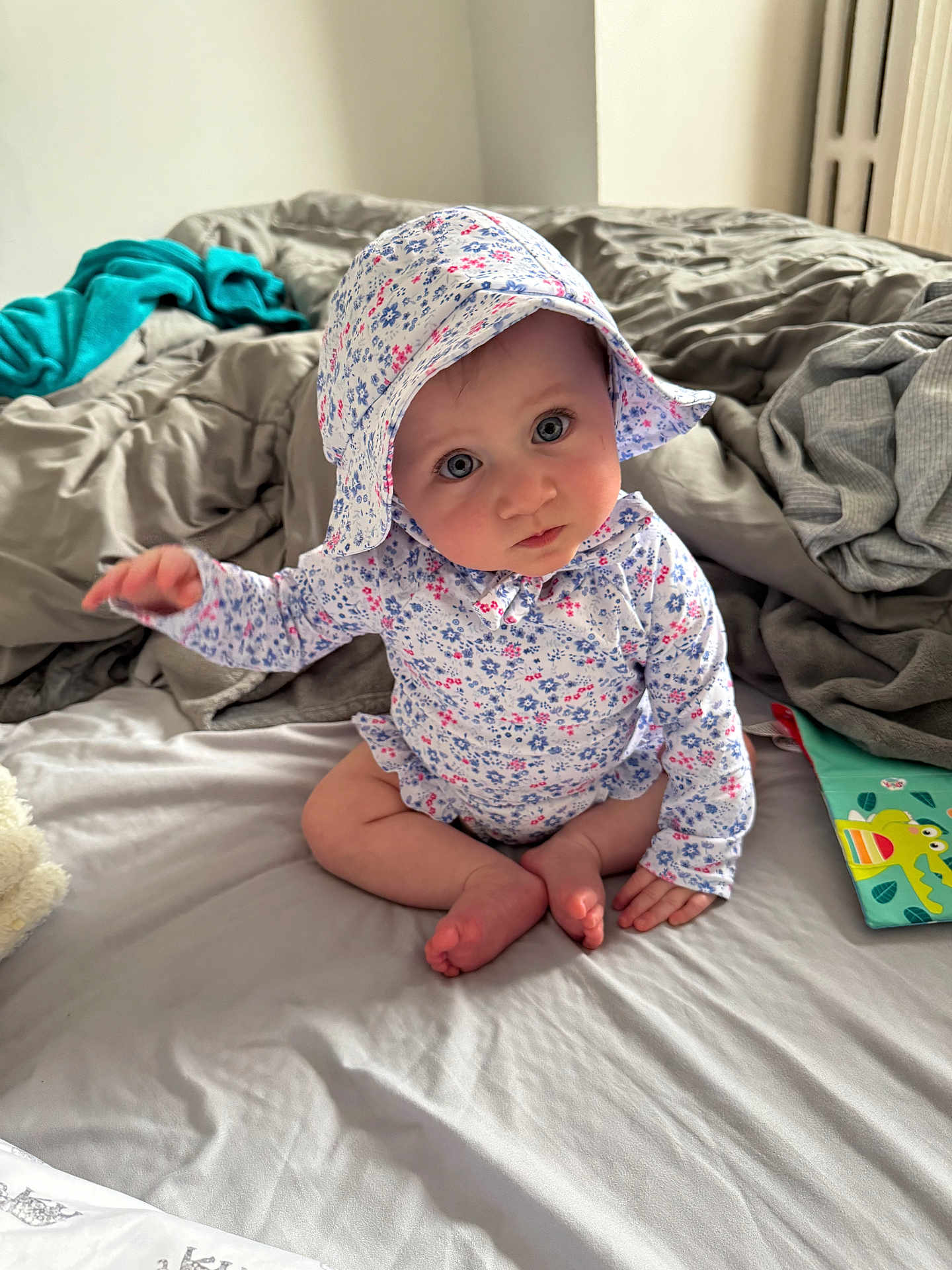 Alessia joined the competition — help win amazing prizes! baby, infant, child, bed, blanket, hat, floral_print, onesie, barefoot, feet, sitting, portrait, big_eyes, blue_eyes, toy_book, bedroom, cozy, cute, curious_gaze, soft_light