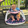 baby, walker, toy, outdoor, porch, wooden_floor, greenery, sunlight, smiling, cheerful, child, infant, plush_toy, clothing, seat, playtime, nature, fence, happy, cute