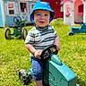 toddler, child, toy, tractor, blue_hat, striped_shirt, outdoor, grass, playhouse, smiling, barefoot, sunny, green, fun, daytime, young_child, happy, summer, play, portrait