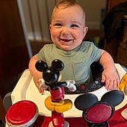 Cassius is registered to the contest to win money with this photo: baby, child, smiling, highchair, toy, mickey_mouse, indoor, happy, cute, infant, face, clothing, person, play, walker, furniture, red, yellow, black, green
