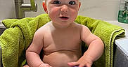 Cassius is registered to the contest to win money with this photo: baby, bath_time, child, clean, curly_hair, cute, expression, indoors, kitchen, person, playful, purple_cup, sink, sitting, skin, tap, towel, water, window, young