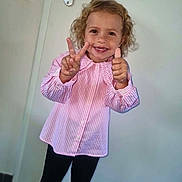 Elina a rejoint le concours — aidez-le/la à gagner de superbes lots ! black_pants, casual_clothing, child, curly_hair, cute, door, expression, full_body, happy, indoor, light_background, peace_sign, person, pink_shirt, portrait, smile, standing, striped_shirt, thumbs_up, young_child