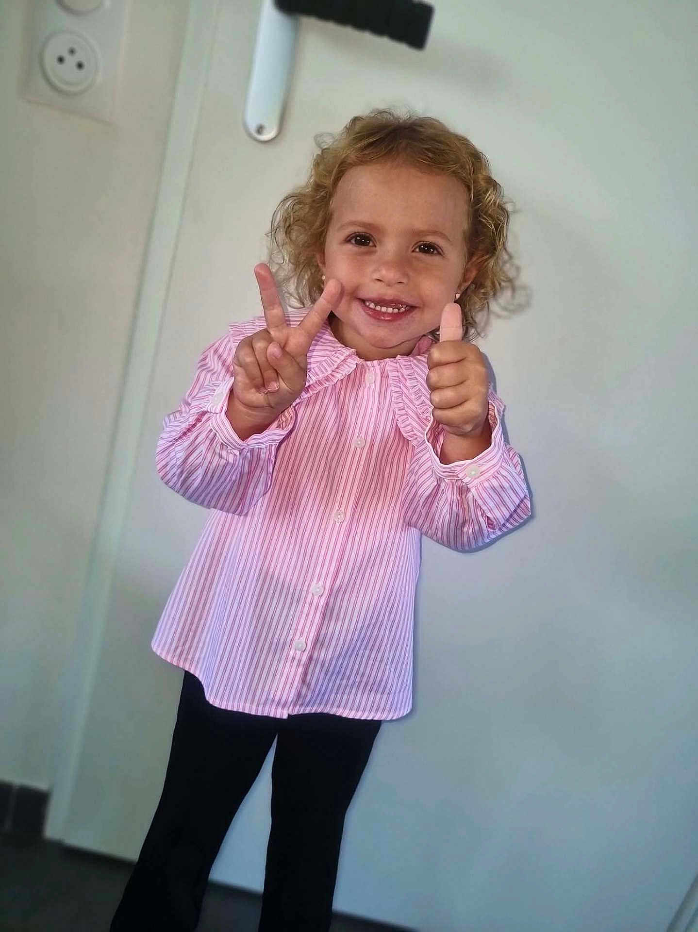 Elina a rejoint le concours — aidez-le/la à gagner de superbes lots ! black_pants, casual_clothing, child, curly_hair, cute, door, expression, full_body, happy, indoor, light_background, peace_sign, person, pink_shirt, portrait, smile, standing, striped_shirt, thumbs_up, young_child