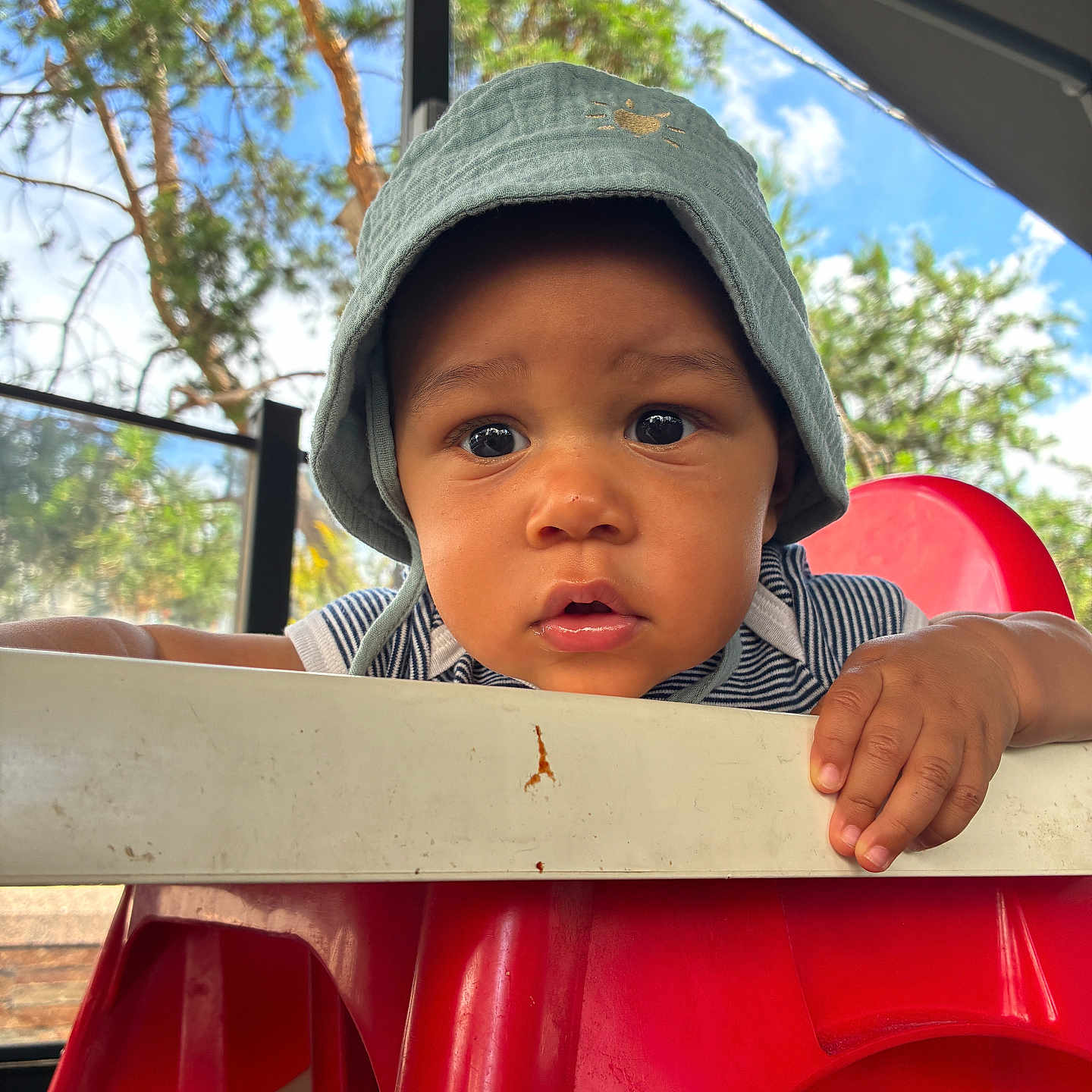Wassym a rejoint le concours — aidez-le/la à gagner de superbes lots ! baby, bucket_hat, child, clothing, curious, daylight, expression, eyes, face, hand, hat, high_chair, outdoor, person, portrait, red_chair, sky, striped_shirt, toddler, tree
