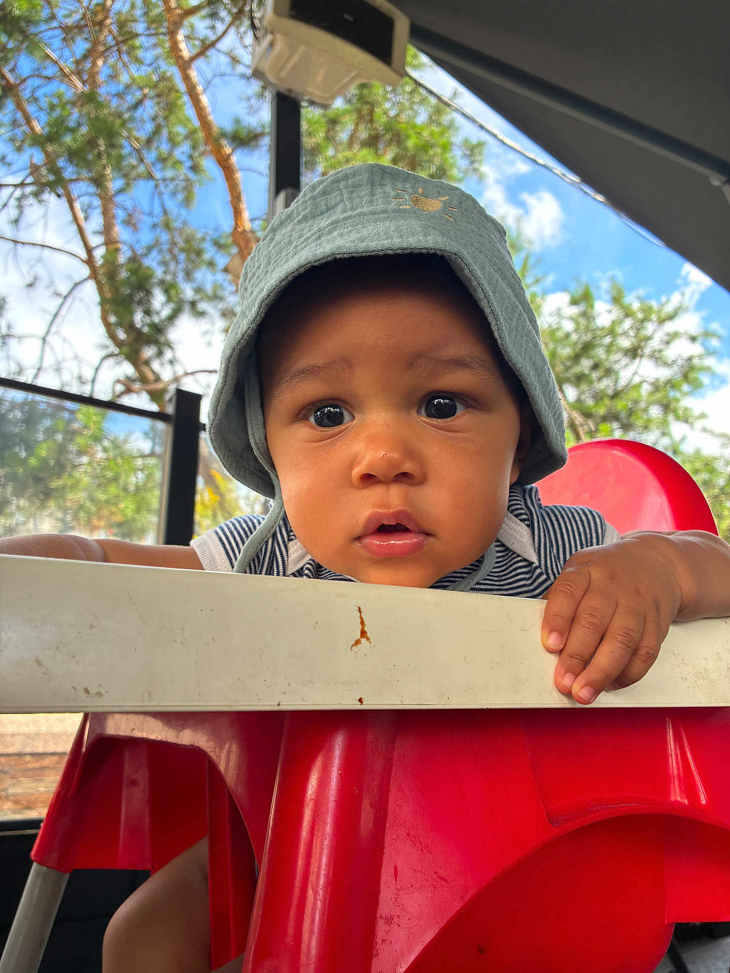 Wassym a rejoint le concours — aidez-le/la à gagner de superbes lots ! toddler, child, baby, hat, bucket_hat, high_chair, red_chair, hand, face, eyes, outdoor, sky, tree, curious, expression, person, portrait, daylight, clothing, striped_shirt