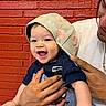 Maclovio is registered to the contest to win money with this photo: adult, baby, brick_wall, cap, child, closeup, clothing, face, family, flowered_hat, happy, hat, holding, indoor, infant, parent, photography, portrait, smiling, spiderman