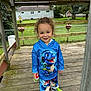 child, toddler, smile, curly_hair, blue_hoodie, dinosaur_graphic, matching_pants, green_sneakers, sneakers, wooden_deck, porch, backyard, chain_link_fence, birdhouse, flower_pot, grass, trees, daylight, portrait, playful