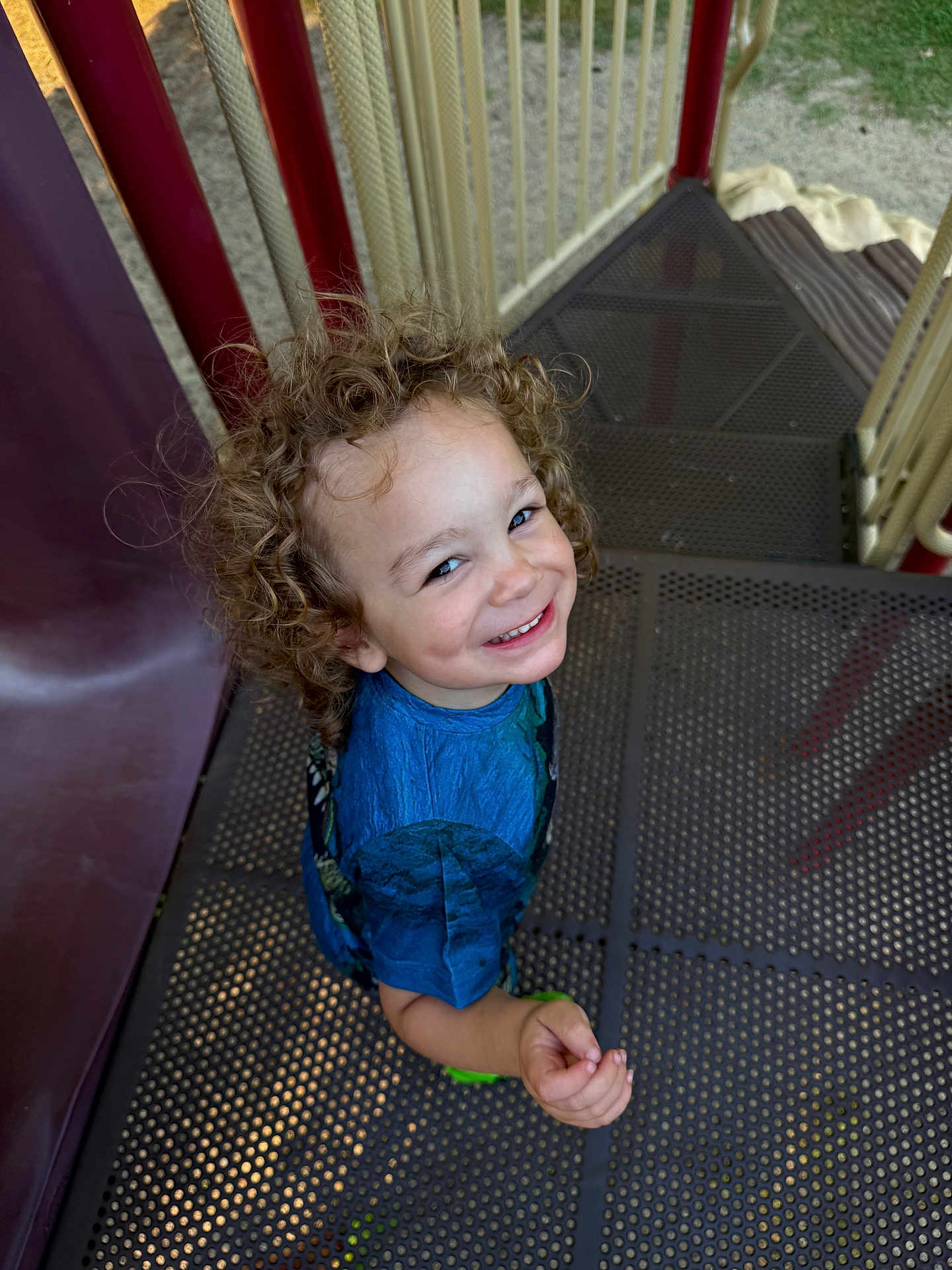 Caspian joined the competition — help win amazing prizes! child, curly_hair, smiling, playground, slide, metal_grating, railing, blue_shirt, looking_up, portrait, happy, toddler, outdoor, cheerful, stairs, play_equipment, candid, sand_ground, green_shoes, person