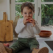 Paul a rejoint le concours — aidez-le/la à gagner de superbes lots ! boots, casual, child, countertop, cutting_board, eating, green_pants, greenery, happy, indoor, kitchen, natural_light, peach, portrait, pottery, sitting, smiling, toddler, white_shirt, window