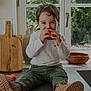 boots, casual, child, countertop, cutting_board, eating, green_pants, greenery, happy, indoor, kitchen, natural_light, peach, portrait, pottery, sitting, smiling, toddler, white_shirt, window