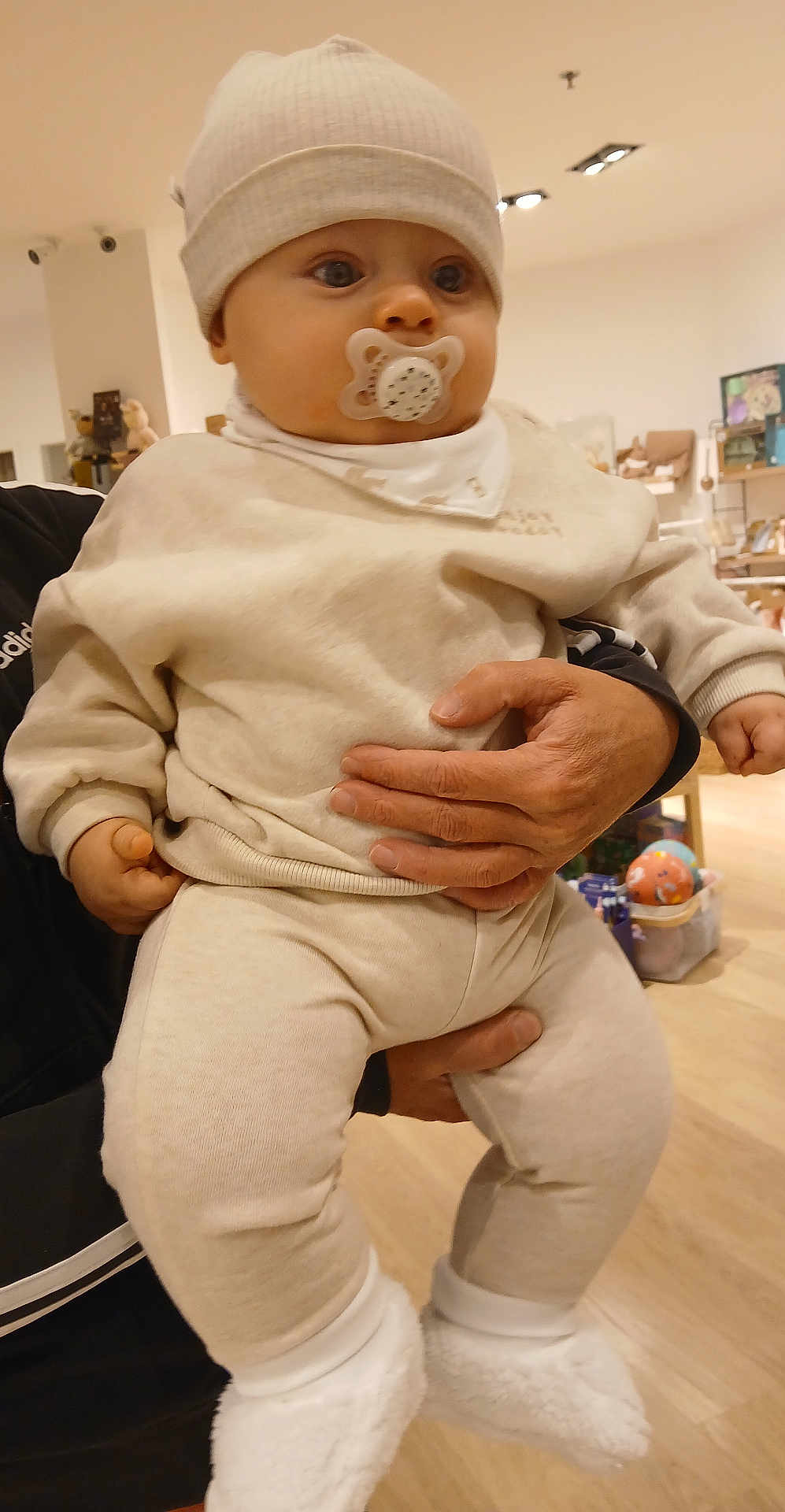 Nino a rejoint le concours — aidez-le/la à gagner de superbes lots ! baby, pacifier, hat, beige_clothing, adult_hand, indoor, toy, shelf, floor, curious, child, footwear, warm_clothing, infant, person, holding, soft_lighting, cute, toddler, furniture