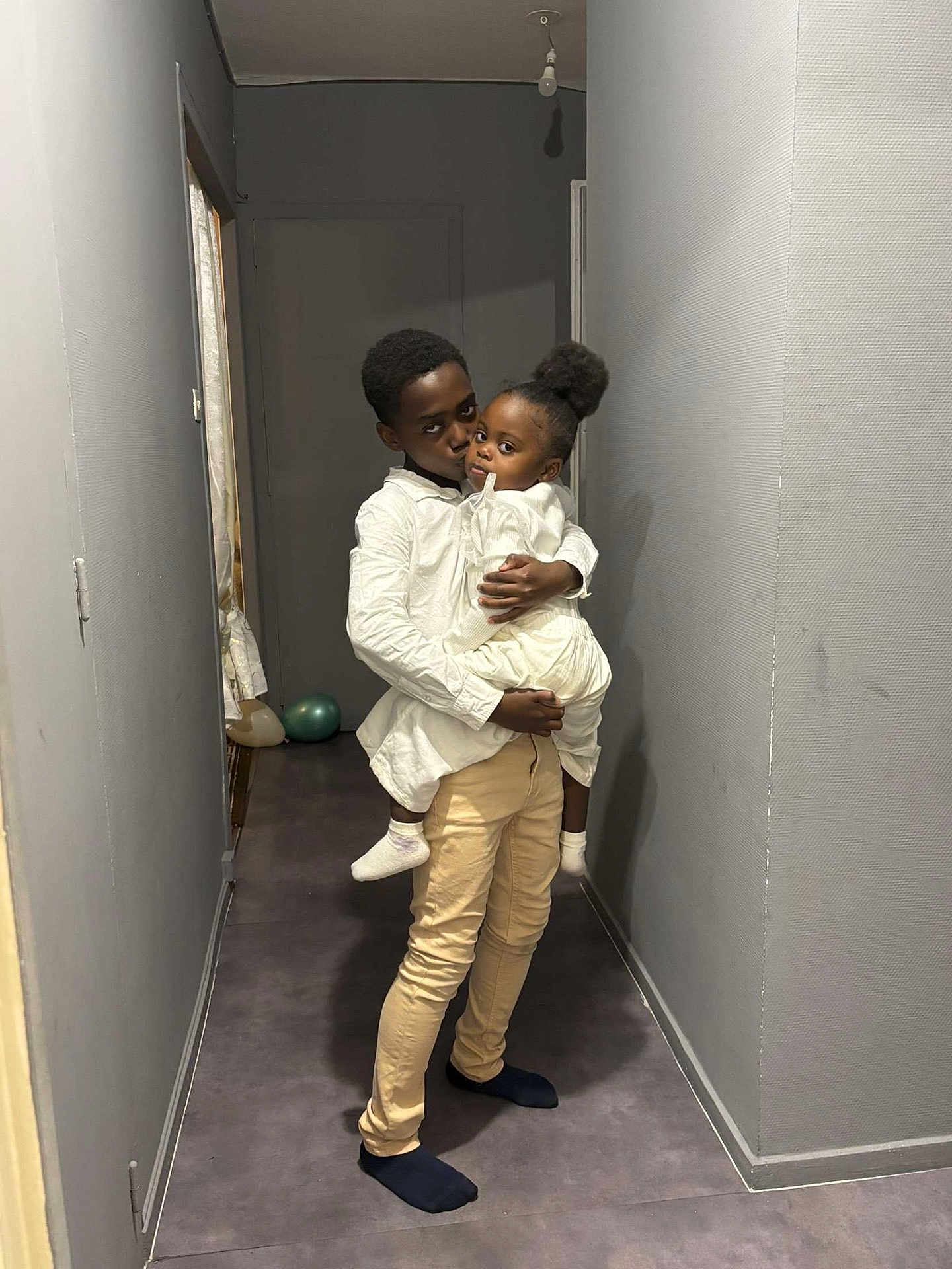 Nelya a rejoint le concours — aidez-le/la à gagner de superbes lots ! child, siblings, hallway, indoor, holding, affection, family, love, clothing, white_shirt, pants, socks, balloons, gray_wall, floor, portrait, people, toddler, boy, girl