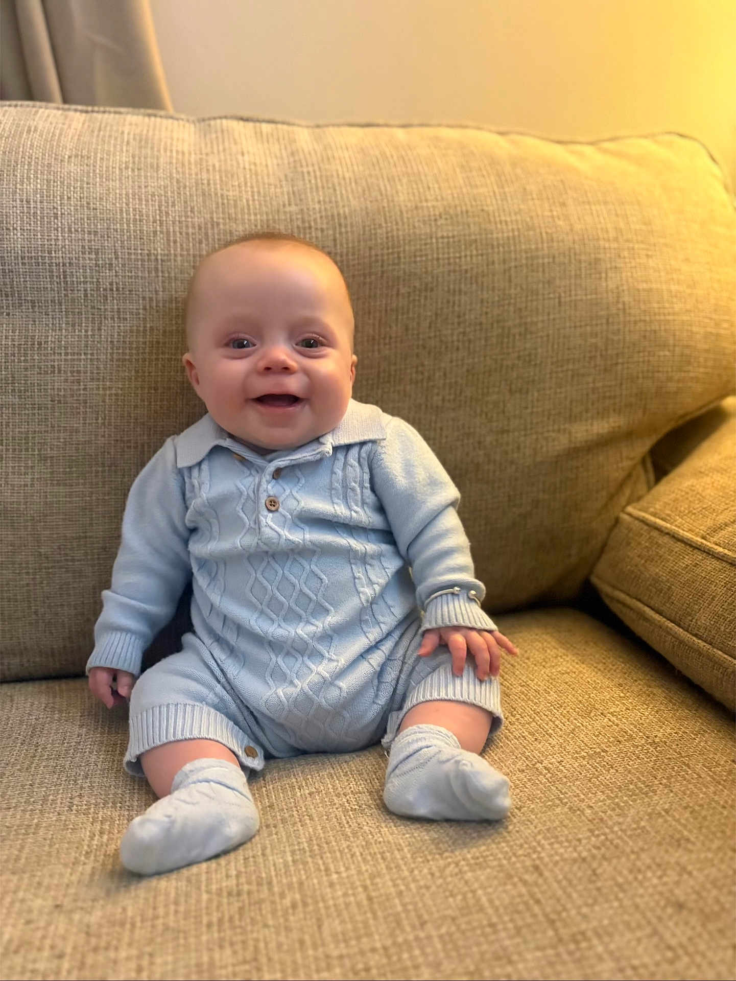 Oakley joined the competition — help win amazing prizes! baby, infant, smile, blue_clothing, knitted_outfit, socks, couch, beige, texture, seated, happy, child, home_interior, portrait, cute, small_hands, small_feet, indoor, warm_lighting, cozy