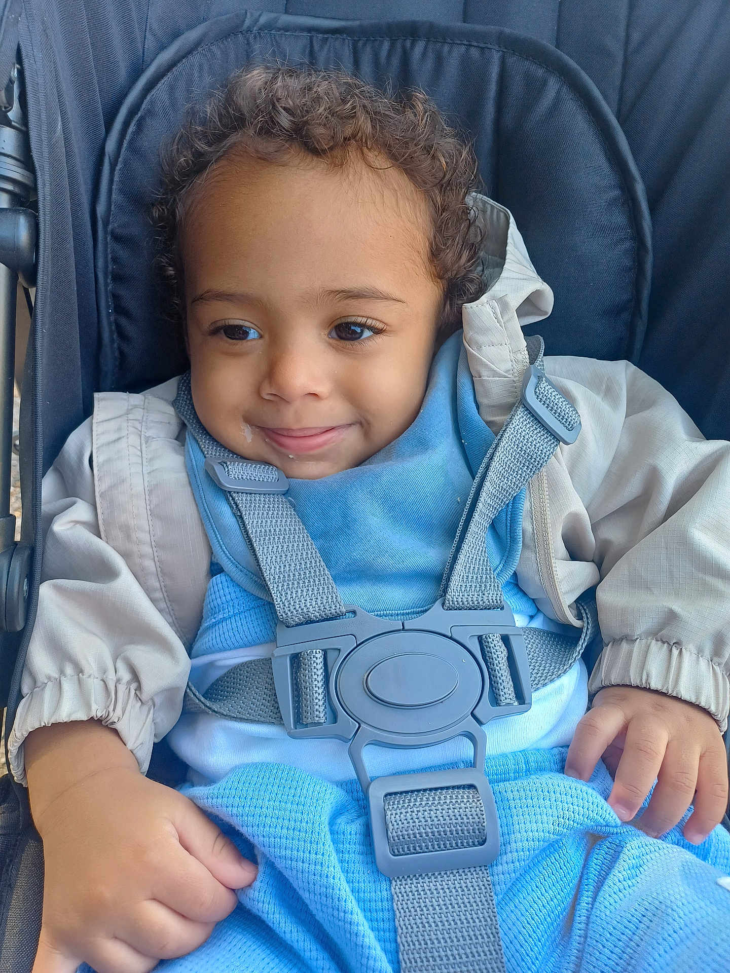 Charlie joined the competition — help win amazing prizes! toddler, child, stroller, curly_hair, smile, blue_clothing, beige_jacket, straps, seatbelt, cute, happy, baby, young_child, portrait, outdoor, casual, hand, face, person, infant