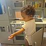 appliance, cabinets, carpet, child, child_playing, curiosity, curly_hair, home, indoor, knobs, open_door, oven, plastic_toys, playset, sink, stove, toddler, toy_kitchen, white_onesie, young_child