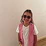 child, girl, sunglasses, pink_vest, white_dress, necklace, backpack, sandals, smiling, standing, wooden_floor, wall, photos, family_photos, indoor, casual, happy, accessories, portrait, fashion