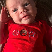 Kaizer is registered to the contest to win money with this photo: baby, infant, red_clothing, face, hand, eyes, smile, lying_down, fabric, pattern, portrait, cute, child, sleepwear, newborn, human, skin, expression, indoors, soft_light