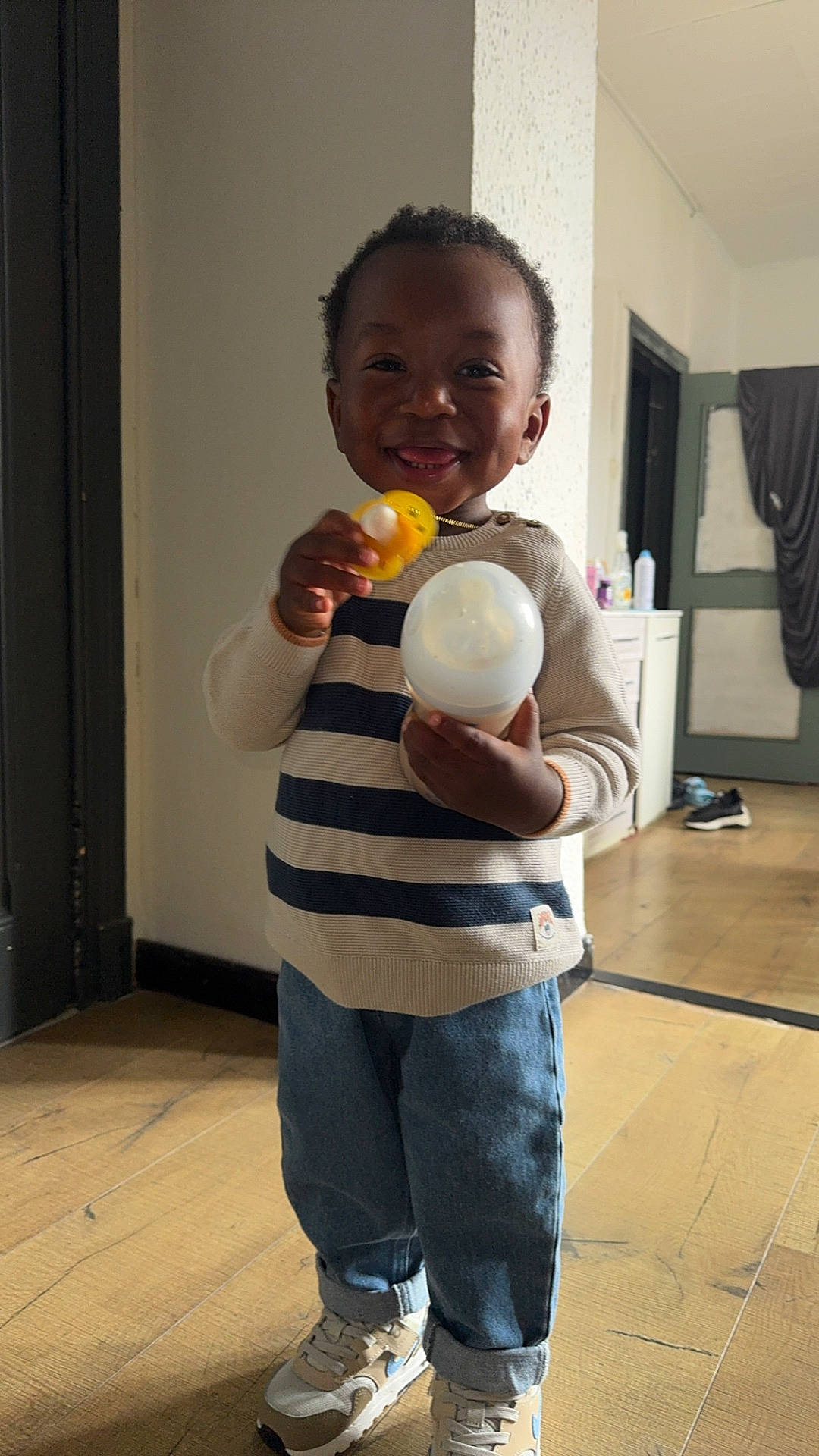 baby_bottle, cabinet, casual_clothing, child, door, face, floor, hair, happy, indoor, jeans, light, person, room, smile, sneakers, standing, striped_sweater, toddler, toy