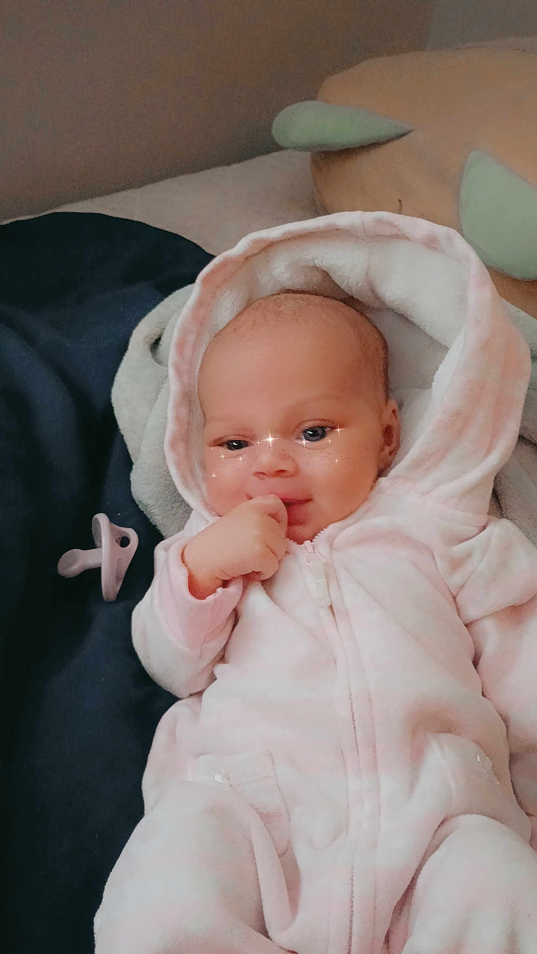 Zanayah is registered to the contest to win money with this photo: baby, infant, pink_onesie, hood, pacifier, blanket, face, sparkle_filter, hand, lying_down, soft_lighting, cute, child, sleepwear, indoors, portrait, expression, skin, head, comfort