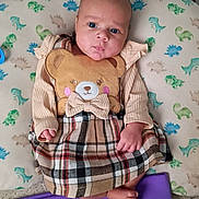 Zanayah is registered to the contest to win money with this photo: baby, infant, child, face, blue_eyes, teddy_bear, bear_applique, plaid_dress, pillow, dinosaur_print, cereal_box, cookie_crisp, bottle, purple_blanket, tiny_feet, hands, indoors, laying_down, cute, clothing