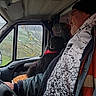 bandana, black_dog, calm, casual_clothing, daytime, dog, glasses, hat, indoors, jacket, leaves, man, portrait, rain, rainy_weather, seat, side_view, tree_branches, vehicle_interior, window