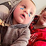 Timothy is registered to the contest to win money with this photo: baby, child, infant, face, portrait, indoor, hoodie, gray_clothing, red_clothing, butterfly, face_paint, smiling, wide_eyes, family, person, close_up, casual, seated, home, photography